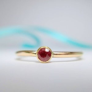 May include: A gold ring with a round, deep red gemstone. The ring has a thin band and a simple, elegant design. The gemstone is set in a bezel setting, and the ring is displayed on a white surface.