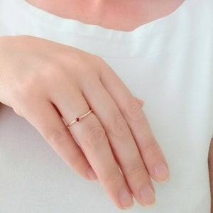 Pu&ograve; includere: Un delicato anello d'oro con una piccola gemma rossa e rotonda al centro. L'anello &egrave; semplice ed elegante, con una fascia sottile. L'anello &egrave; su un dito, sullo sfondo di una camicia bianca.