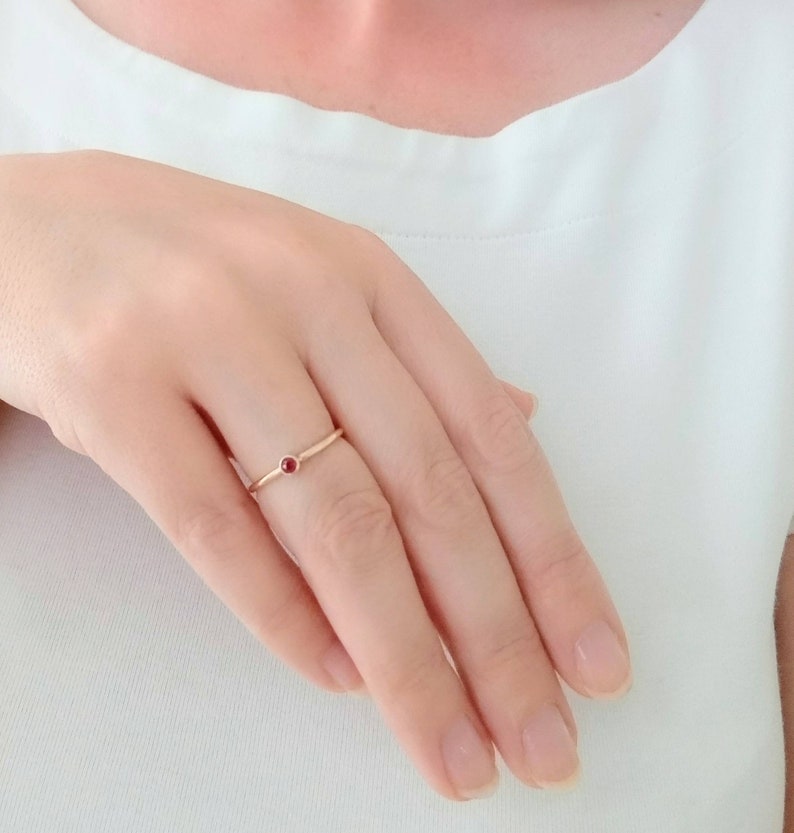 Pu&ograve; includere: Un delicato anello d'oro con una piccola gemma rossa e rotonda al centro. L'anello &egrave; semplice ed elegante, con una fascia sottile. L'anello &egrave; su un dito, sullo sfondo di una camicia bianca.