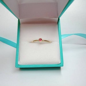 Pu&ograve; includere: Un delicato anello d'oro con una piccola gemma rossa e rotonda &egrave; esposto all'interno di un cofanetto turchese con interno bianco. Un nastro turchese &egrave; drappeggiato attorno alla scatola. L'anello &egrave; centrato nella scatola.