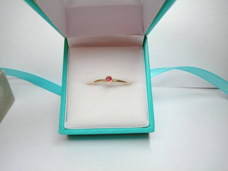 Pu&ograve; includere: Un delicato anello d'oro con una piccola gemma rossa e rotonda &egrave; esposto all'interno di un cofanetto turchese con interno bianco. Un nastro turchese &egrave; drappeggiato attorno alla scatola. L'anello &egrave; centrato nella scatola.