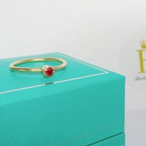 Pu&ograve; includere: Un delicato anello d'oro con una piccola gemma rossa e rotonda. L'anello &egrave; posto su una scatola turchese, su sfondo bianco. L'anello ha un design semplice ed elegante, perfetto per l'uso quotidiano. Il logo "Beebee Jewellery" &egrave; visibile.