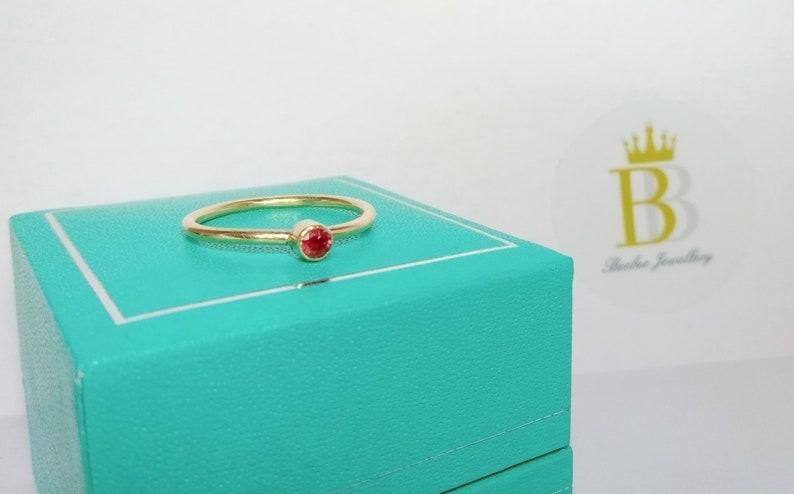 Pu&ograve; includere: Un delicato anello d'oro con una piccola gemma rossa e rotonda. L'anello &egrave; posto su una scatola turchese, su sfondo bianco. L'anello ha un design semplice ed elegante, perfetto per l'uso quotidiano. Il logo "Beebee Jewellery" &egrave; visibile.