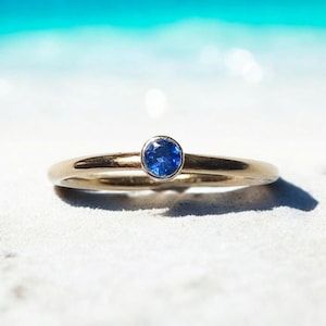 May include: A delicate gold band ring with a single, small, round, blue sapphire gemstone.