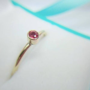 Pu&ograve; includere: Un delicato anello d'oro con una piccola gemma rotonda color rubino. L'anello ha una fascia sottile e un design semplice. La gemma &egrave; incastonata in una lunetta, evidenziando il suo colore vibrante contro l'oro.