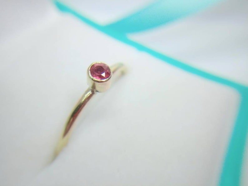 Pu&ograve; includere: Un delicato anello d'oro con una piccola gemma rotonda color rubino. L'anello ha una fascia sottile e un design semplice. La gemma &egrave; incastonata in una lunetta, evidenziando il suo colore vibrante contro l'oro.