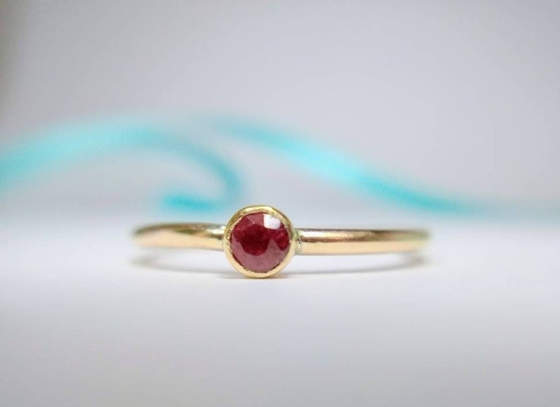 Pu&ograve; includere: Un anello d'oro con una gemma rotonda di colore rosso intenso. L'anello ha una fascia sottile e un design semplice ed elegante. La gemma &egrave; incastonata in una lunetta, su una superficie bianca.