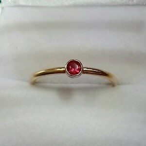 Pu&ograve; includere: Un delicato anello d'oro con una piccola gemma rossa vibrante e rotonda. L'anello ha una fascia sottile e un design semplice ed elegante. La gemma &egrave; incastonata in una lunetta, esaltando il suo colore e la sua brillantezza.