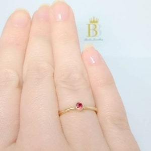 Pu&ograve; includere: Un delicato anello d'oro con una piccola gemma rossa e rotonda. L'anello &egrave; mostrato su un dito, evidenziando il suo design semplice ma elegante. La fascia lucida mette in risalto il colore vibrante della gemma.