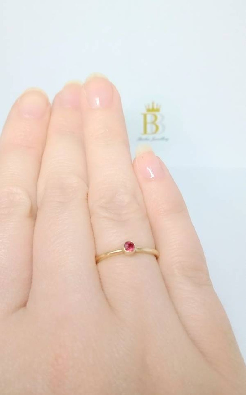 Pu&ograve; includere: Un delicato anello d'oro con una piccola gemma rossa e rotonda. L'anello &egrave; mostrato su un dito, evidenziando il suo design semplice ma elegante. La fascia lucida mette in risalto il colore vibrante della gemma.