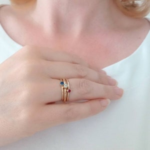 Pu&ograve; includere: Una mano che indossa tre anelli d'oro. Ogni anello presenta una gemma diversa: uno zaffiro blu, un diamante trasparente e un rubino rosso. Gli anelli sono impilati sul dito, creando un look a strati.