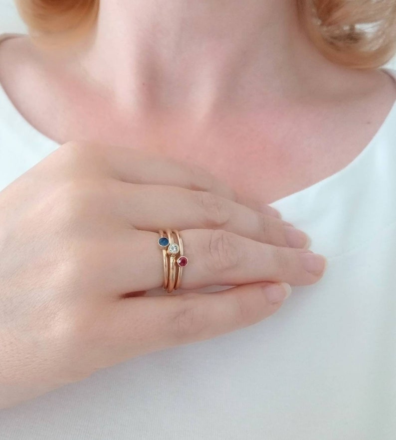 Pu&ograve; includere: Una mano che indossa tre anelli d'oro. Ogni anello presenta una gemma diversa: uno zaffiro blu, un diamante trasparente e un rubino rosso. Gli anelli sono impilati sul dito, creando un look a strati.