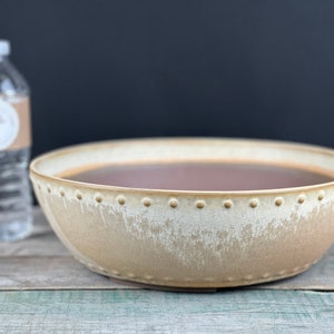 May include: A large, round, light brown ceramic planter with a textured, speckled glaze. The planter has a raised rim with a row of small, evenly spaced dots.