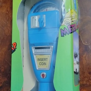 May include: A blue toy parking meter in its original box. The meter has a dial, a coin slot, and a red button. The box is green with the word "METER" in red and a picture of a car.