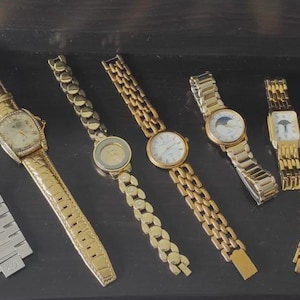 May include: Collection of wristwatches featuring various designs. Includes watches with gold and silver bands, some with decorative faces. One watch has a leather strap. The watches are displayed on a dark surface.