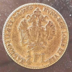 Può includere: Una moneta d'oro con dettagli intricati, con un'aquila a due teste al centro. Il bordo della moneta è inciso con testo e l'anno 1859. La moneta ha anche il testo "1/4 FL".