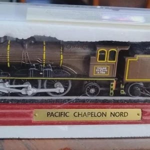 May include: A brown and black model train with yellow detailing, presented in a white box. The train sits on a red base with a gold plaque reading "PACIFIC CHAPELON NORD". The model is a detailed replica.