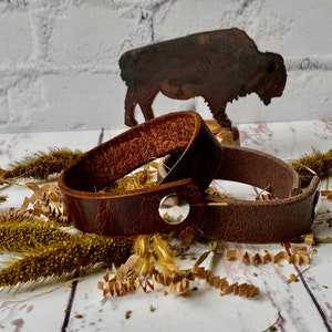 May include: Two brown leather bracelets with a silver snap closure. The bracelets are lying on a white wooden surface with dried grass and brown paper shreds.