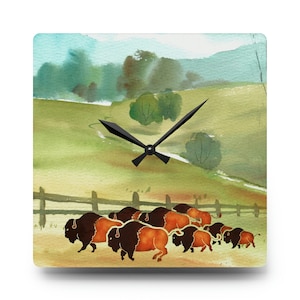 May include: Square wall clock featuring a watercolor painting of a herd of bison in a grassy field. The clock face has a rustic fence, green hills, and trees in the background. Black clock hands indicate the time.