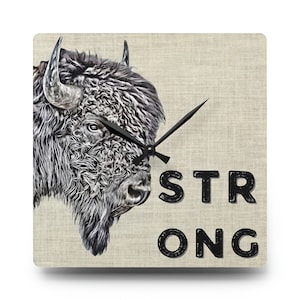 May include: Square wall clock featuring a detailed illustration of a bison's head in shades of gray and white. The clock face has a textured, beige background with the word "STRONG" in black. Black clock hands indicate the time.