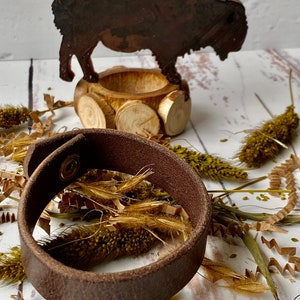 May include: A brown leather bracelet with a metal snap closure. The bracelet is lying on a white surface with dried grasses and a wooden bison figurine.