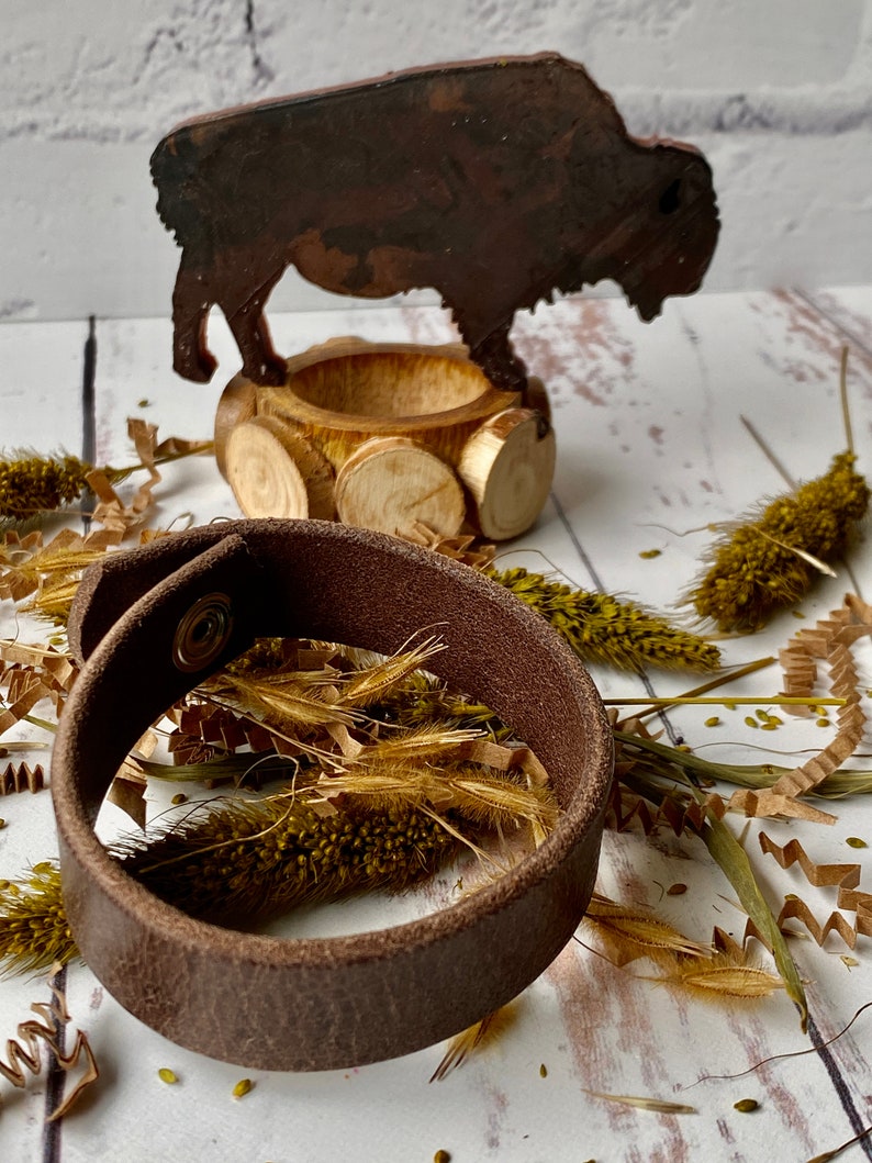 May include: A brown leather bracelet with a metal snap closure. The bracelet is lying on a white surface with dried grasses and a wooden bison figurine.