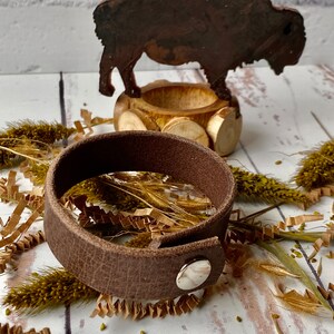 May include: A brown leather bracelet with a silver snap closure. The bracelet is lying on a white surface with brown paper shreds and dried grass.