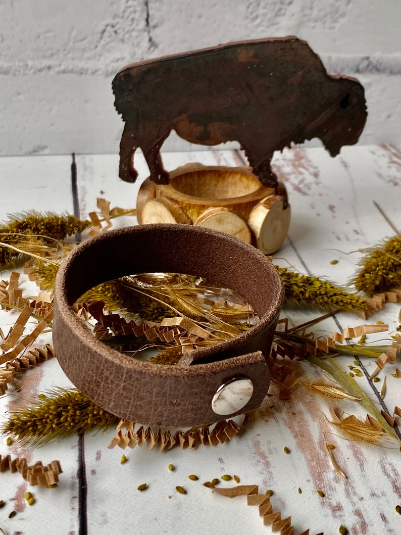 May include: A brown leather bracelet with a silver snap closure. The bracelet is lying on a white surface with brown paper shreds and dried grass.