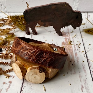 May include: A brown leather bracelet with a wooden base made of stacked wood rounds. The bracelet is wrapped around the wood base and has a metal clasp.