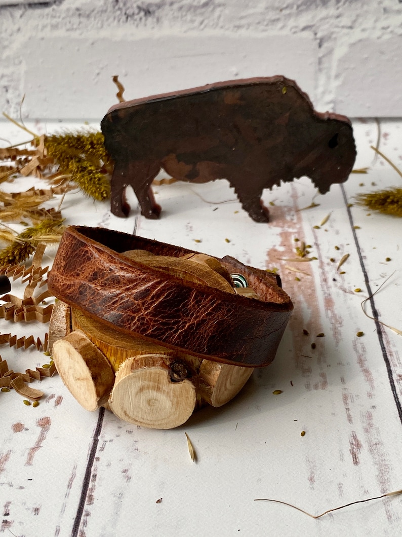 May include: A brown leather bracelet with a wooden base made of stacked wood rounds. The bracelet is wrapped around the wood base and has a metal clasp.