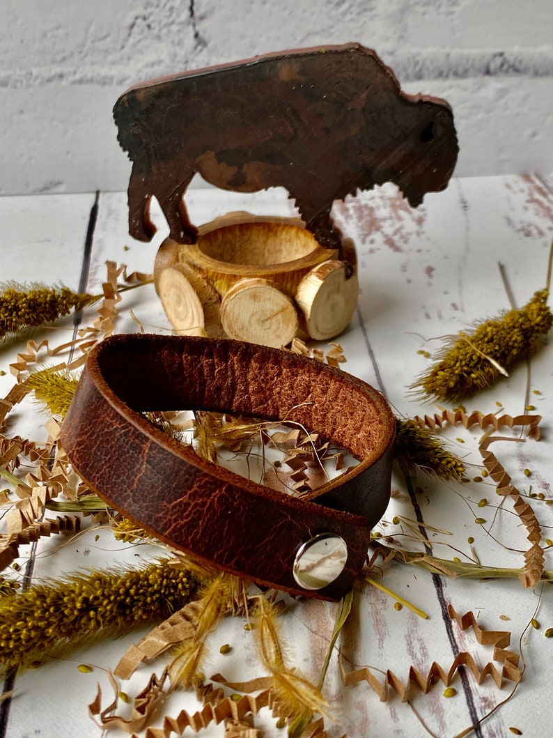 May include: A brown leather bracelet with a silver snap closure. The bracelet is lying on a white surface with dried grasses and paper shreds.