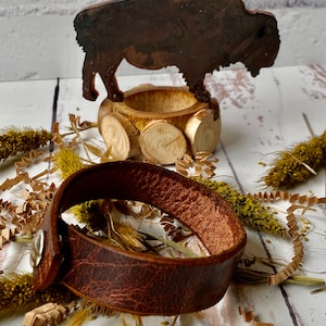 May include: A brown leather bracelet with a silver snap closure. The bracelet is lying on a white surface with brown paper shreds and dried flowers.