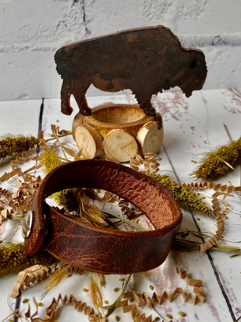 May include: A brown leather bracelet with a silver snap closure. The bracelet is lying on a white surface with brown paper shreds and dried flowers.
