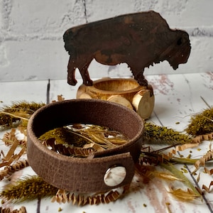 May include: A brown leather bracelet with a silver snap closure. The bracelet is lying on a white surface with dried grass and paper shreds.