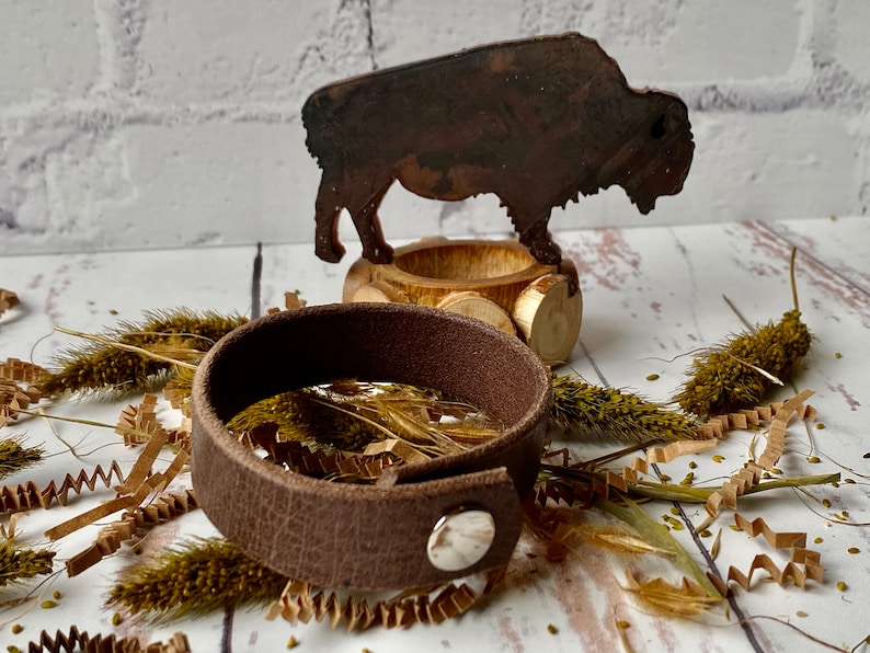 May include: A brown leather bracelet with a silver snap closure. The bracelet is lying on a white surface with dried grass and paper shreds.