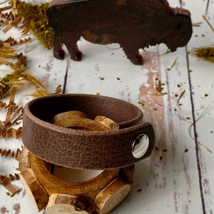 May include: A brown leather bracelet with a silver snap closure and a wooden ring with a natural finish.