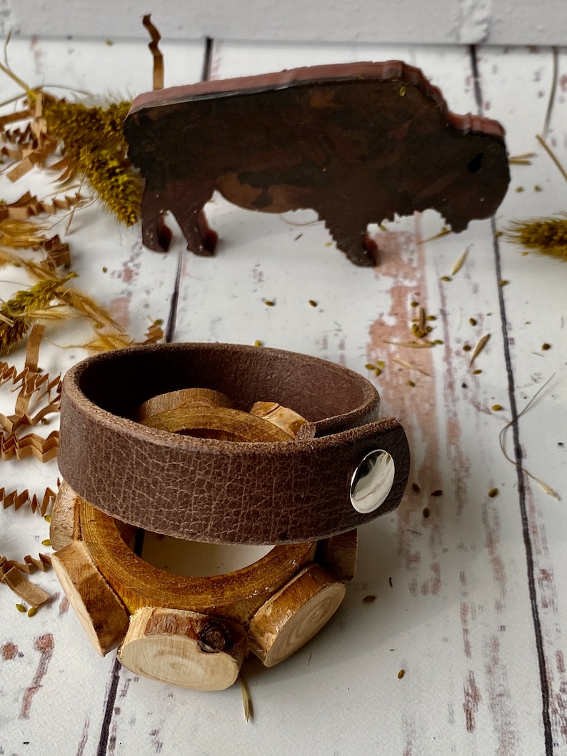 May include: A brown leather bracelet with a silver snap closure and a wooden ring with a natural finish.