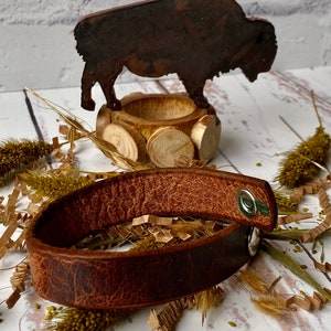 May include: A brown leather bracelet with a silver snap closure. The bracelet is lying on a white surface with brown paper shreds and dried grasses.