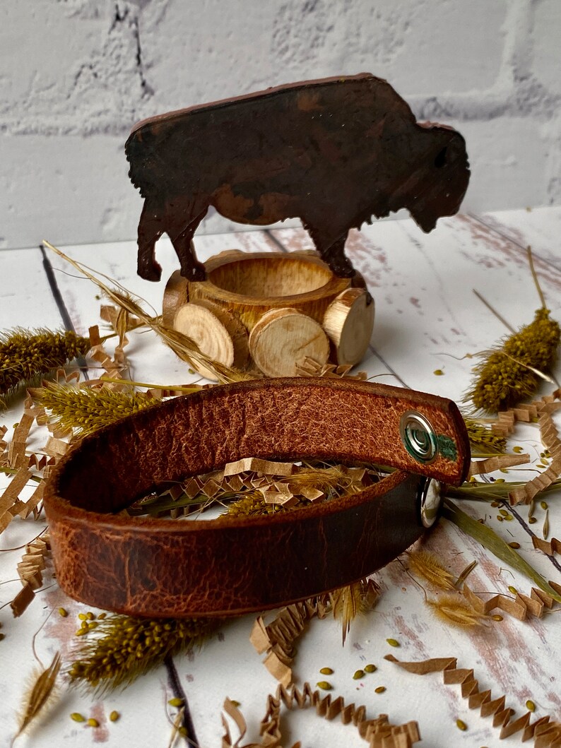May include: A brown leather bracelet with a silver snap closure. The bracelet is lying on a white surface with brown paper shreds and dried grasses.