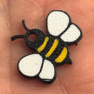 May include: A small, decorative bee-shaped embellishment. The bee features a black body with yellow stripes and white wings. The wings have a textured appearance. The bee is a craft supply item.