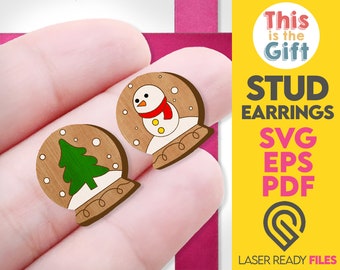 May include: A pair of wooden stud earrings with a Christmas theme. One earring features a snowman with a red scarf and the other earring features a green Christmas tree inside a snow globe.
