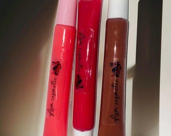 Pode incluir: Três tubos de brilho labial dispostos verticalmente. O tubo da esquerda é rosa brilhante com tampa rosa, o central é vermelho escuro com tampa branca e o da direita é castanho com brilhos dourados e tampa branca. Cada tubo tem um rótulo transparente com o texto Asm cosmetic.