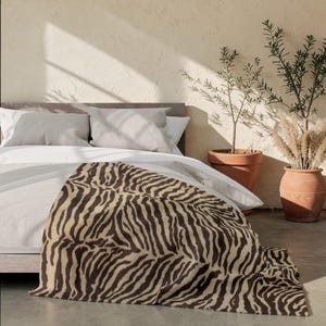May include: A soft throw blanket featuring a dark brown and cream zebra stripe pattern draped over a bed with white bedding and two light gray pillows. The blanket displays a repeating animal print design across its entire visible surface area.