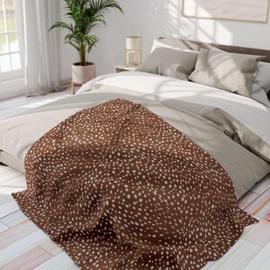 May include: A brown throw blanket featuring a repeating pattern of small, irregular white dots draped over a bed. The fabric has a soft texture and covers the lower portion of the bedding, contrasting with the neutral-toned pillows and sheets.