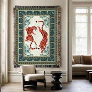 May include: A rectangular woven tapestry featuring two orange tigers with black stripes against a cream background. The tapestry has a teal border with floral and geometric patterns, and fringed edges. The artwork is displayed on a wall.