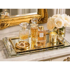 May include: A mirrored tray with gold trim holds perfume bottles, a bracelet, and a small dish with pearls. A lit candle in a gold holder and a bouquet of white roses are also on the tray. The background includes a gold-framed mirror.