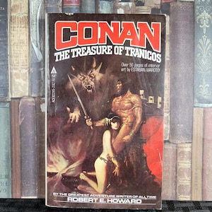 Könnte beinhalten: Ein Taschenbuchcover mit einer roten und weißen Illustration von Conan dem Barbaren, einer Frau und einem Wolf. Der Buchtitel lautet "Conan The Treasure of Tranicos" mit dem Text "Over 50 pages of interior art by ESTABAN MAROTO" unter dem Titel. Das Buch ist von Robert E. Howard.