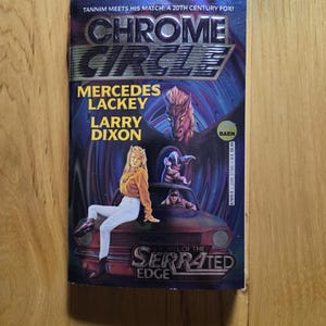以下が含まれることがあります： メルセデス・ラッキーとラリー・ディクソンによる「Chrome Circle: A Novel of the Serrated Edge」というタイトルのペーパーバック。表紙には、白いズボンとオレンジ色のトップスを着た女性が車の上に座っており、背景にはドラゴンや他の人物が描かれています。
