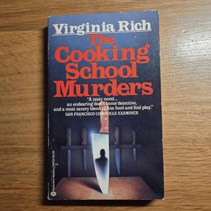 May include: A paperback book titled The Cooking School Murders by Virginia Rich. The dark blue cover features a large chef knife with a silhouette of a person reflected in the blade, positioned in front of a rack of knives and a wooden surface.
