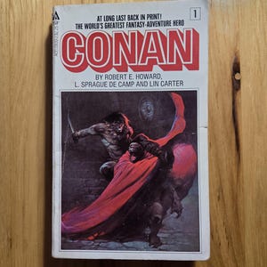May include: A vintage paperback book titled "CONAN" with a cover illustration of a warrior in a red cape battling a monster. The book is number 1 in the series and features text that reads "AT LONG LAST BACK IN PRINT!"